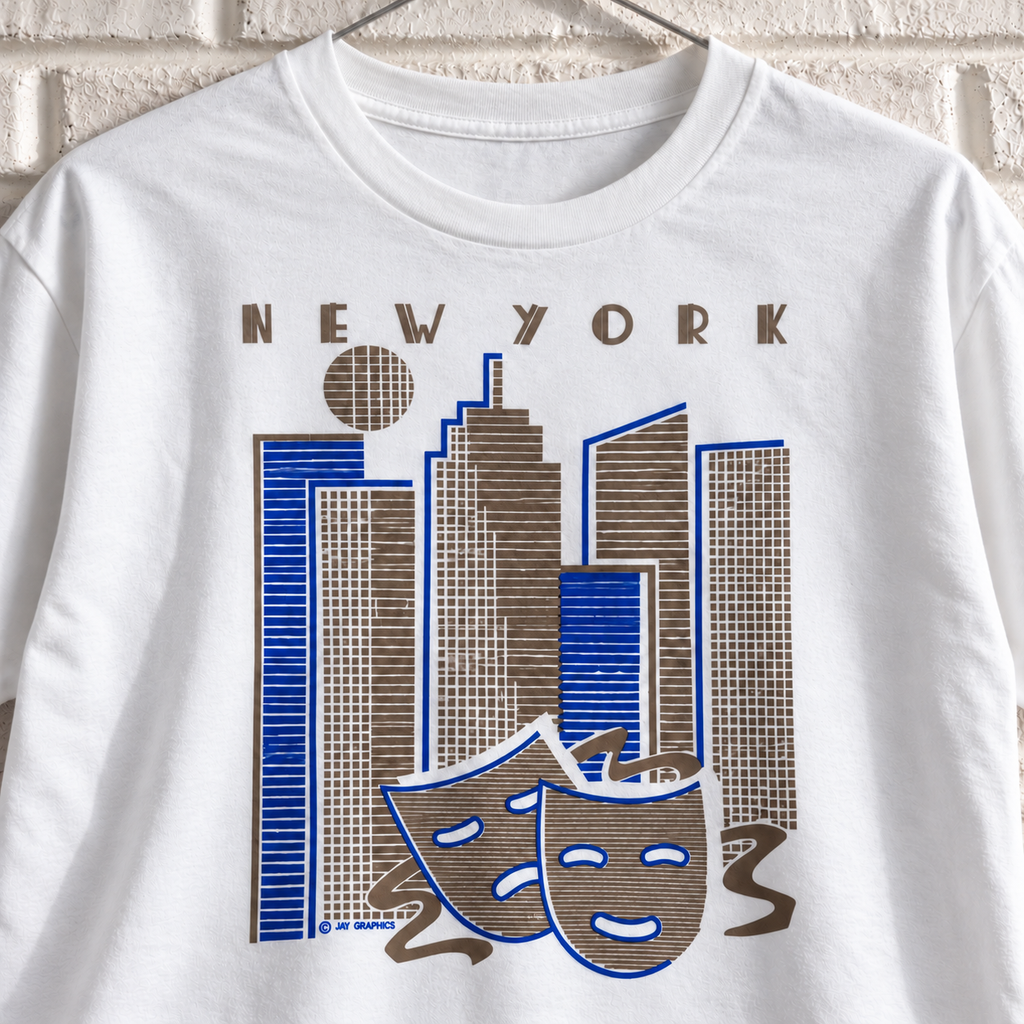 White New York skyline and theatre masks graphic tee on a white brick wall with a wire hanger — close-up front view