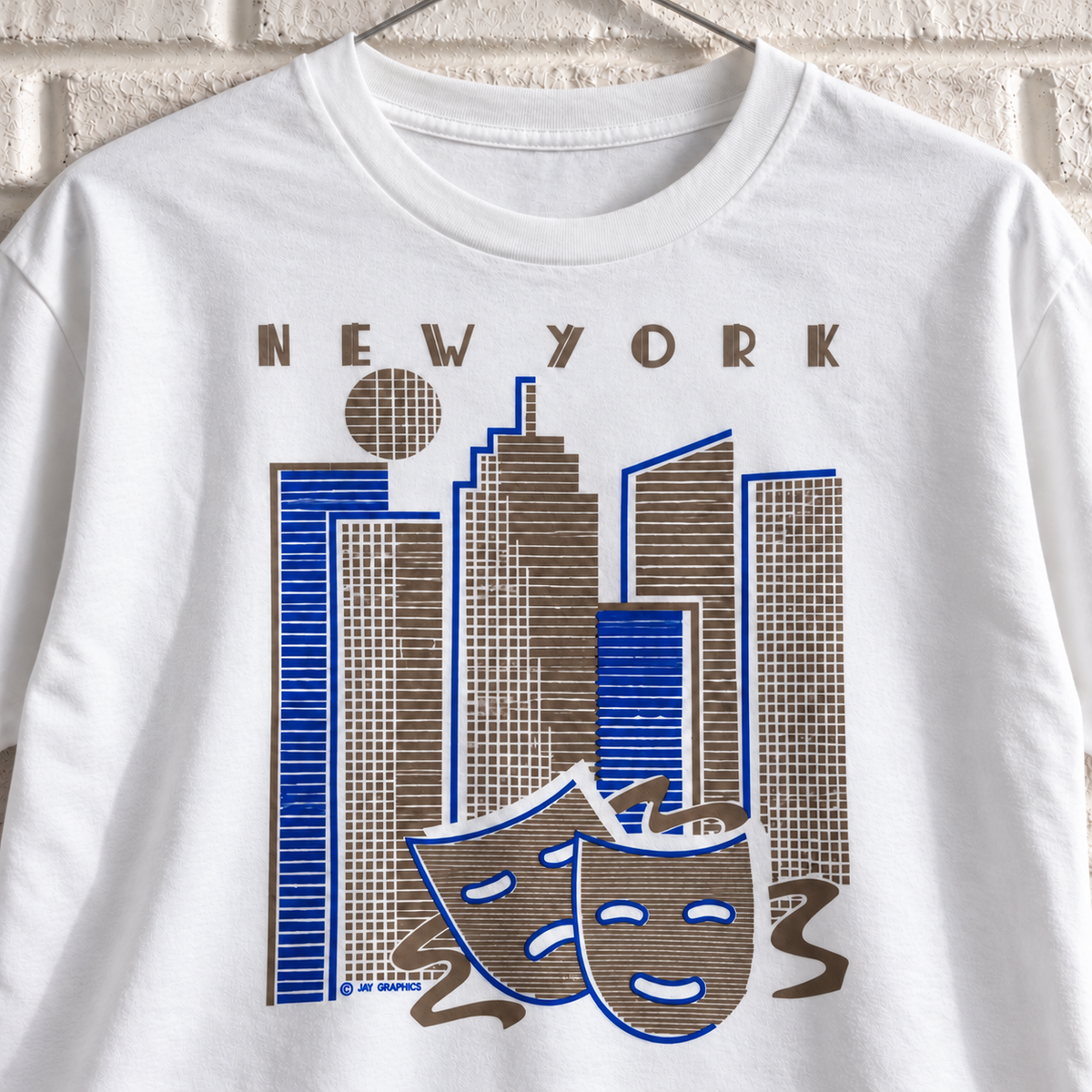 White New York skyline and theatre masks graphic tee on a white brick wall with a wire hanger — close-up front view