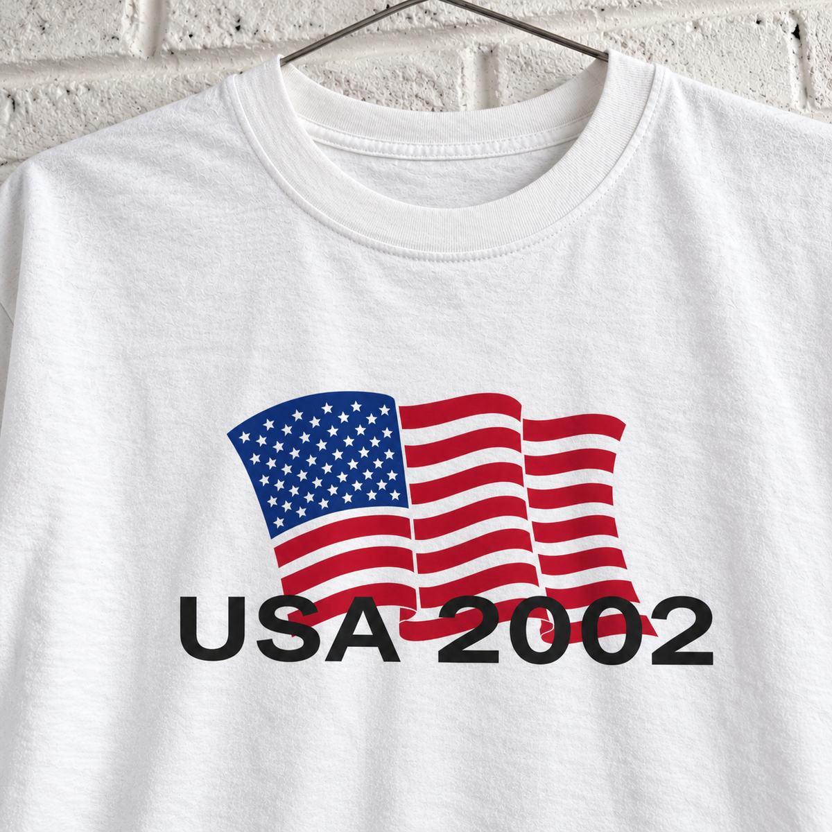 USA 2002 American flag graphic tee on a white brick wall with a wire hanger — close-up front view