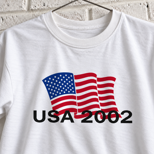 USA 2002 American flag graphic tee on a white brick wall with a wire hanger — close-up front view