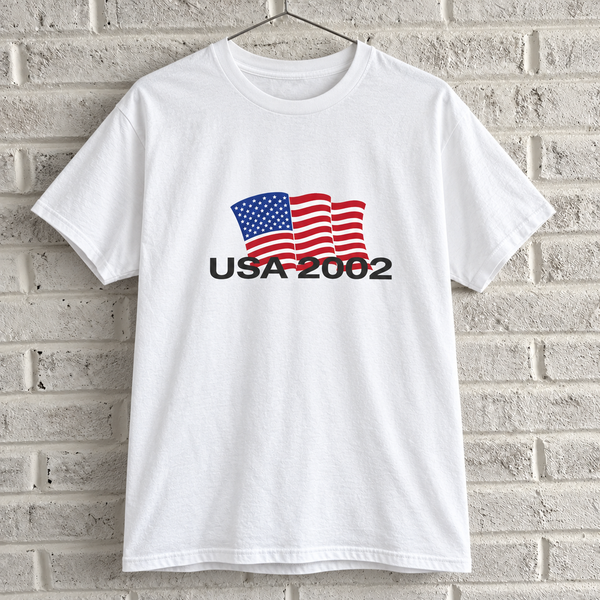 USA 2002 American flag graphic tee on a white brick wall with a wire hanger — front view