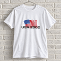 USA 2002 American flag graphic tee on a white brick wall with a wire hanger — front view