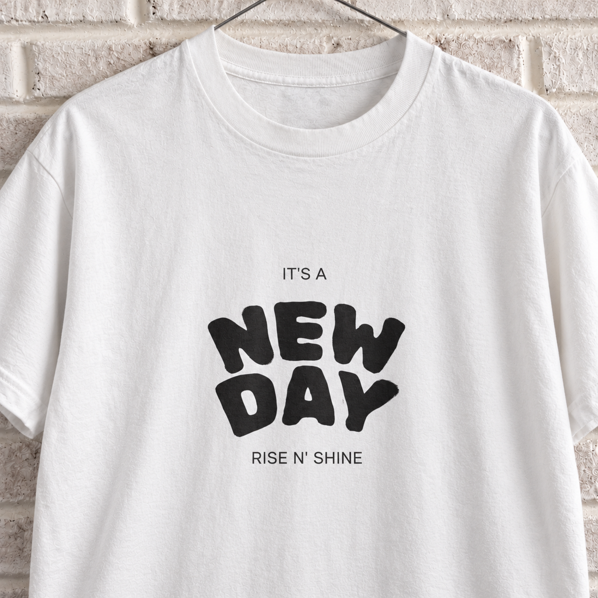 White Comfort Colors graphic tee on a white brick wall with a wire hanger — close-up front view